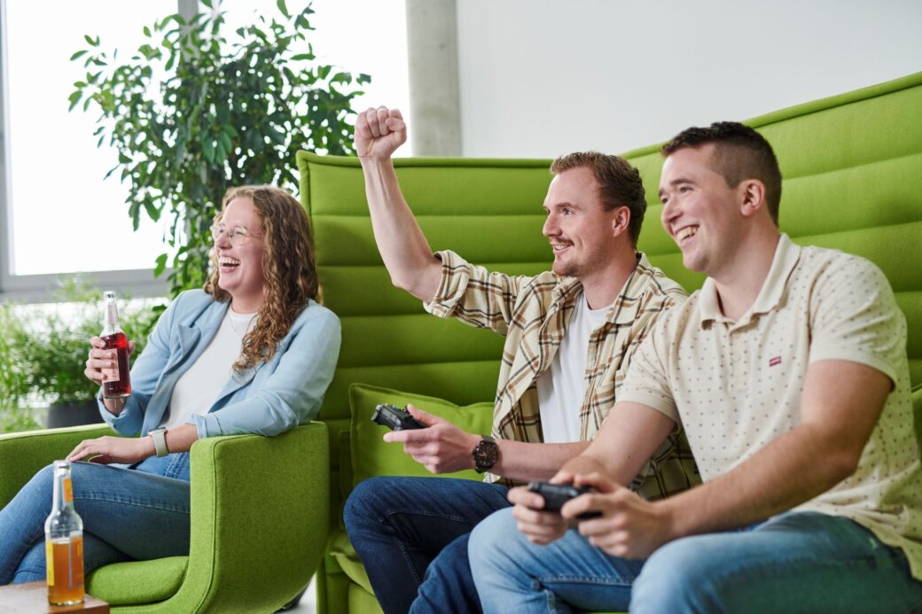 Mitarbeitende sitzen auf einer Couch und spielen gemeinsam an einer Spielekonsole