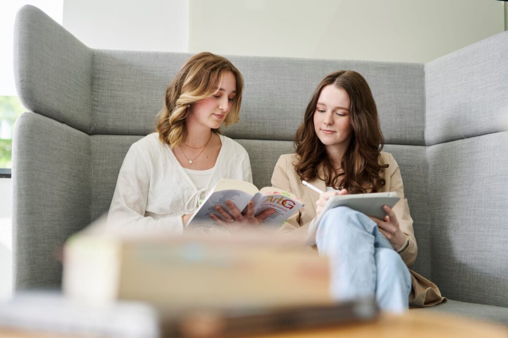 Mitarbeiterinnen sitzten gemeinsam auf einer Couch und schauen in ihre Lernbücher