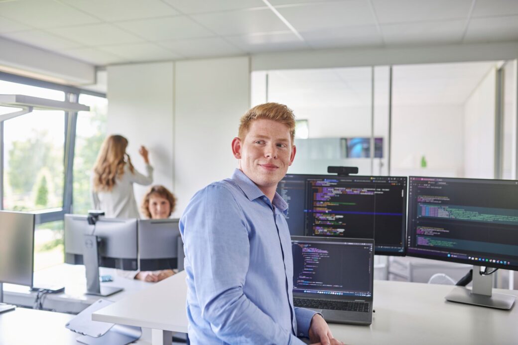 Mitarbeiter steht an seinem Schreibtisch und hat auf seinen Bildschirmen eine Programmieroberfläche geöffnet
