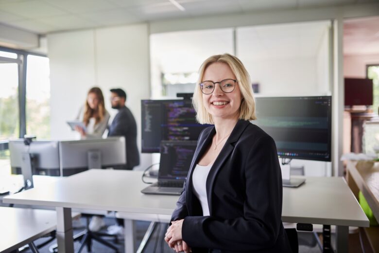 A colleague from the development department stands at her desk and smiles into the camera.